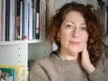 Elisabeth Åsbrink, med blå ögon och lockigt hår, sitter framför en bokhylla fylld med böcker.