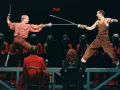 Scen ur Hamlet där två män duellerar med svärd. Bakom dem står figurer i röda dräkter med masker. Vad handlar Hamlet om? Kanske svek och hämnd.