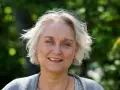 Porträtt av Karin Alvtegen med blont hår och blå ögon, leende utomhus mot en grönskande bakgrund.