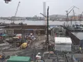 Byggarbetsplats vid vattnet med kranar, grävmaskiner och byggmaterial. Arbetet med att underminera marken pågår.