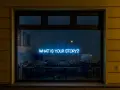 Blå neontext "WHAT IS YOUR STORY?" lyser i ett fönster, som en inbjudan till en skrivarkurs online.