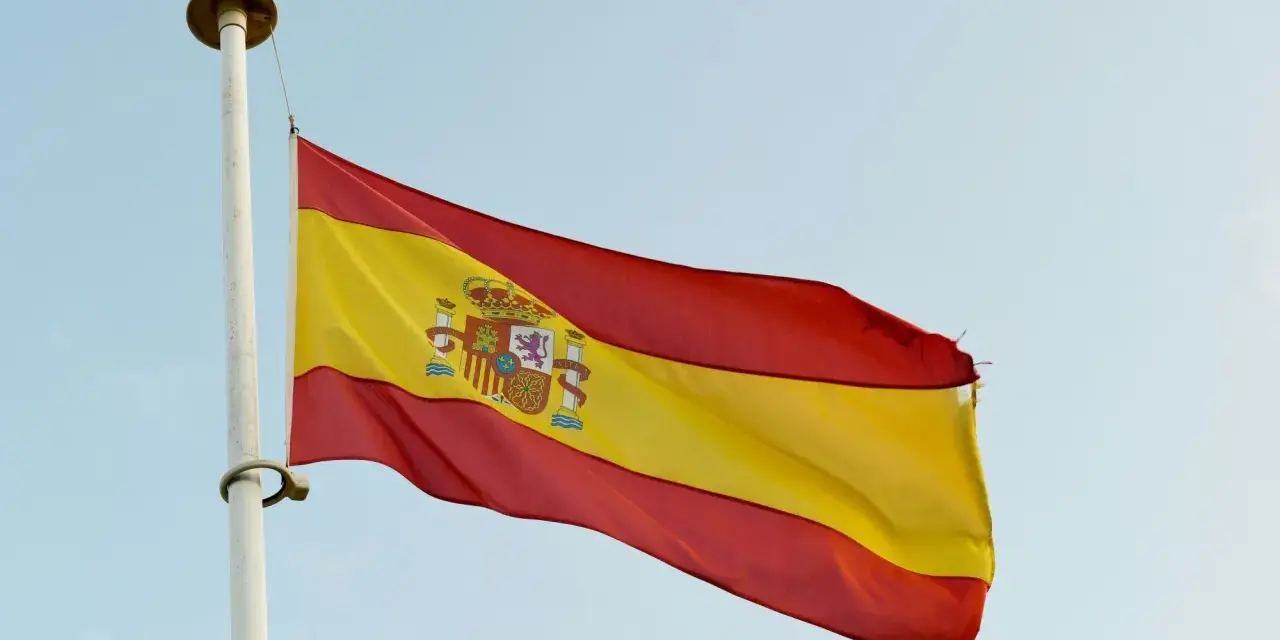 Spanska flaggan vajar i vinden. Den har tre horisontella band: rött, gult och rött. I det gula bandet finns Spaniens riksvapen. Orden "spanska ord" kan associeras med denna flagga.