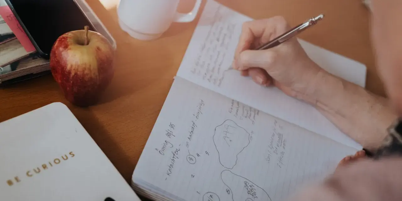 En hand skriver i ett anteckningsblock. Bredvid ligger en mobil, ett äpple och en mugg. Tidningen skriva är en del av en anteckning.