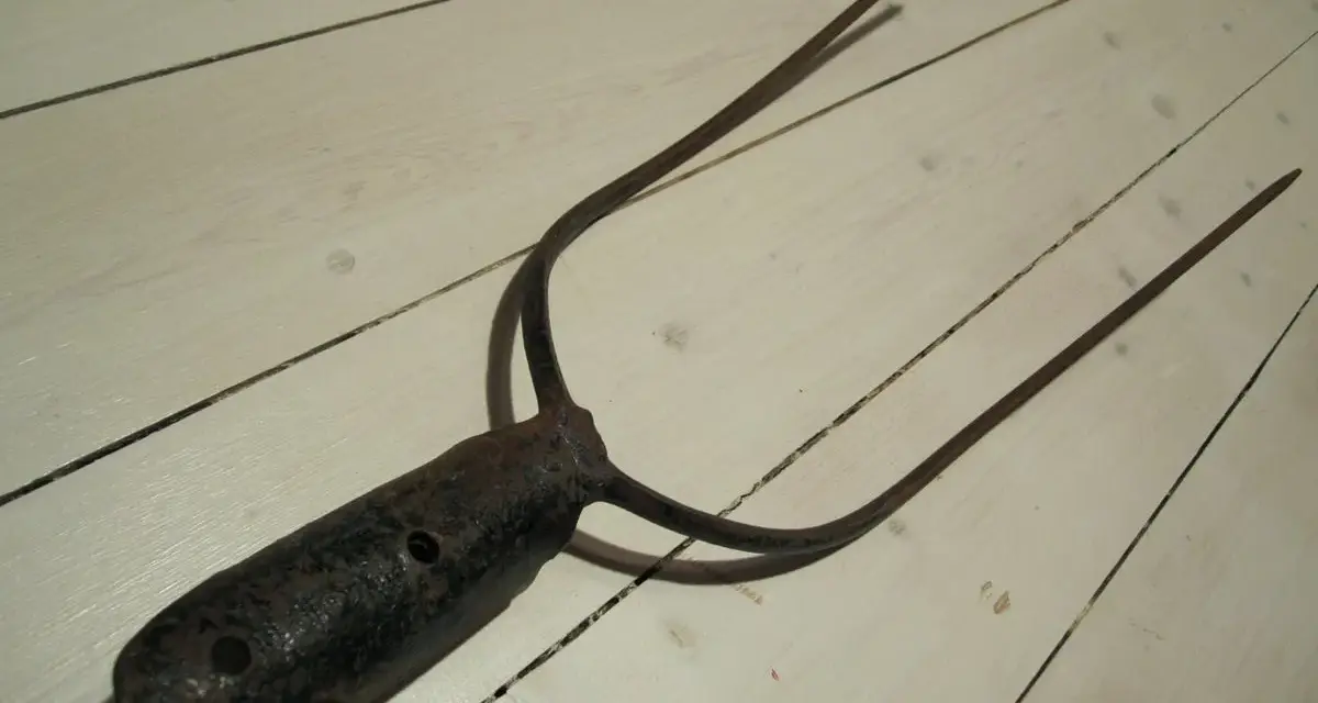 A rusty, old pitchfork with three tines lies on a white wooden floor.