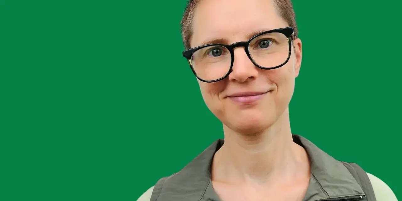 En kvinna med glasögon ler mot kameran. Hon har kortklippt hår och bär en grön väst. Bilden ger informerande text.