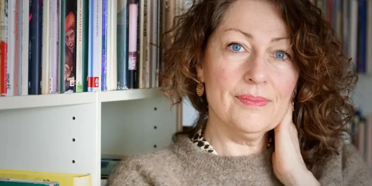 Elisabeth Åsbrink, med blå ögon och lockigt hår, sitter framför en bokhylla fylld med böcker.