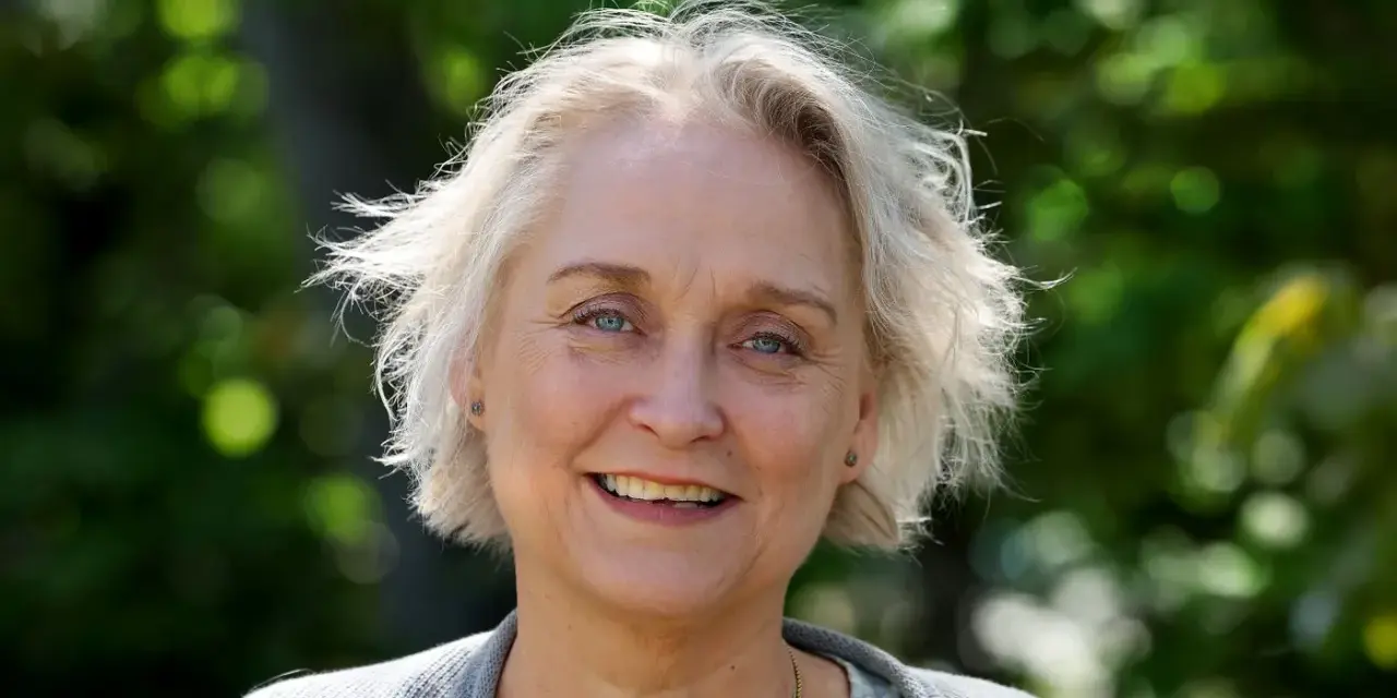 Porträtt av Karin Alvtegen med blont hår och blå ögon, leende utomhus mot en grönskande bakgrund.