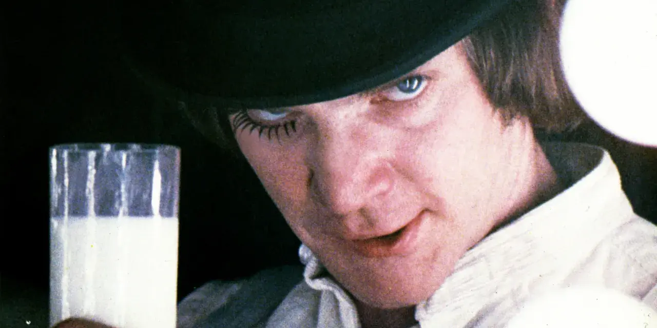 En ung man med hatt och falska ögonfransar håller ett glas mjölk. Vem skrev A Clockwork Orange? Anthony Burgess.