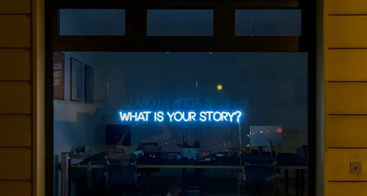 Blå neontext "WHAT IS YOUR STORY?" lyser i ett fönster, som en inbjudan till en skrivarkurs online.