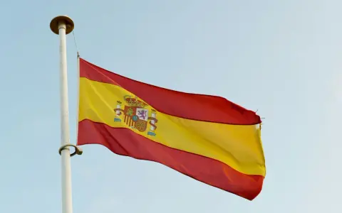 Spanska flaggan vajar i vinden. Den har tre horisontella band: rött, gult och rött. I det gula bandet finns Spaniens riksvapen. Orden "spanska ord" kan associeras med denna flagga.