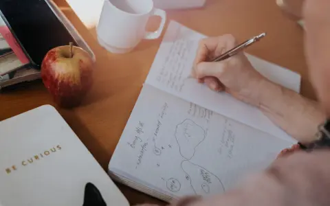 En hand skriver i ett anteckningsblock. Bredvid ligger en mobil, ett äpple och en mugg. Tidningen skriva är en del av en anteckning.