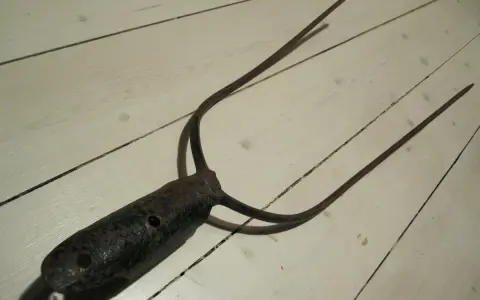 A rusty, old pitchfork with three tines lies on a white wooden floor.
