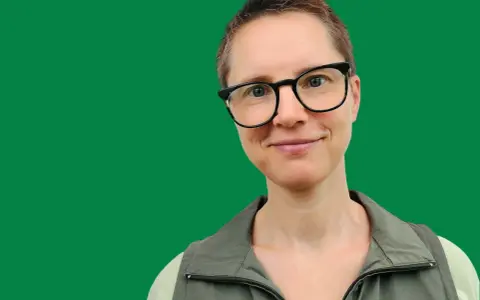 En kvinna med glasögon ler mot kameran. Hon har kortklippt hår och bär en grön väst. Bilden ger informerande text.