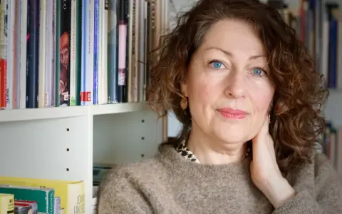 Elisabeth Åsbrink, med blå ögon och lockigt hår, sitter framför en bokhylla fylld med böcker.
