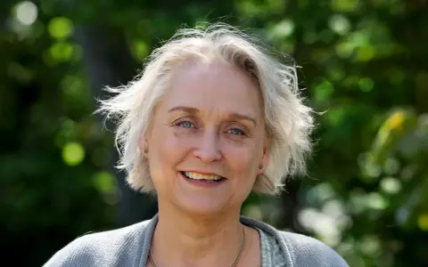 Porträtt av Karin Alvtegen med blont hår och blå ögon, leende utomhus mot en grönskande bakgrund.