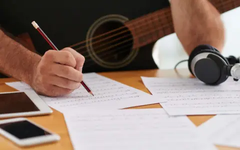 En person skriver noter med en penna, med en gitarr och hörlurar i bakgrunden. Detta är en bra inledning till en ny låt.