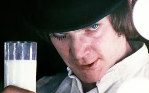 En ung man med hatt och falska ögonfransar håller ett glas mjölk. Vem skrev A Clockwork Orange? Anthony Burgess.