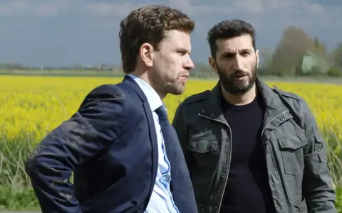 Två män står i en gul rapsåker. Den ena bär kostym, den andra en militärjacka. Scen från avdelning q film.