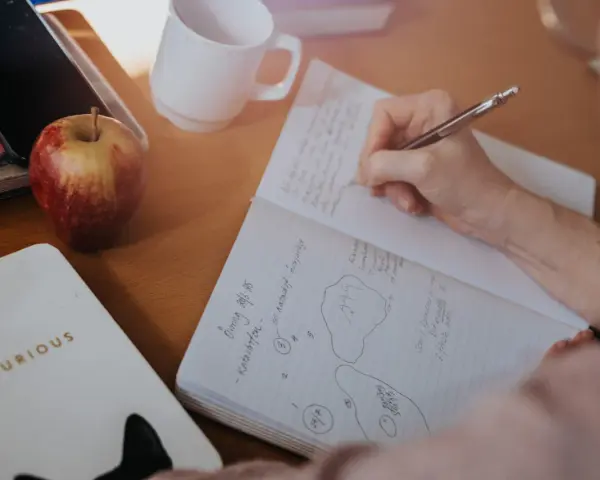 En hand skriver i ett anteckningsblock. Bredvid ligger en mobil, ett äpple och en mugg. Tidningen skriva är en del av en anteckning.