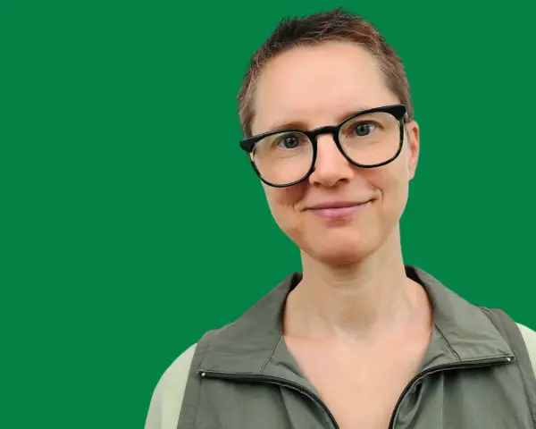 En kvinna med glasögon ler mot kameran. Hon har kortklippt hår och bär en grön väst. Bilden ger informerande text.