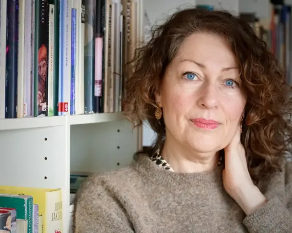 Elisabeth Åsbrink, med blå ögon och lockigt hår, sitter framför en bokhylla fylld med böcker.