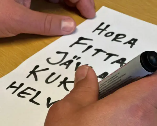 En hand skriver en svordomar lista på ett papper. Orden "hora", "fitta", "jäv", "kuk" och "helv" syns tydligt.