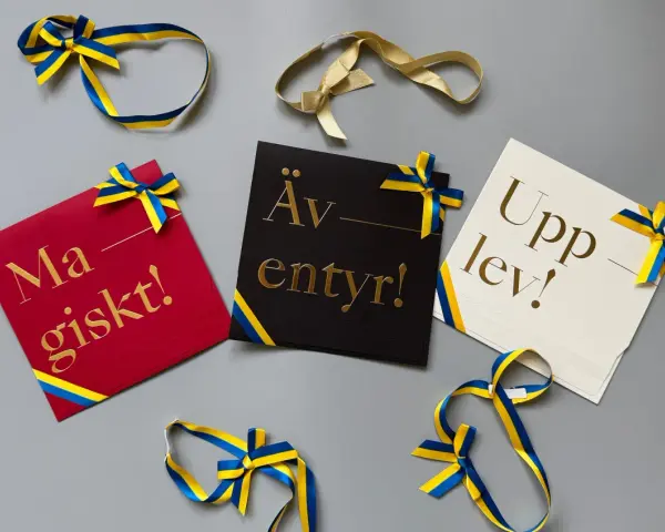 Grattis! Kort med texten "Magiskt!", "Äventyr!" och "Upplev!" i guldfärgad skrivstil, dekorerade med blågula band.