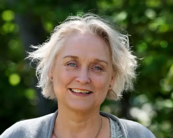 Porträtt av Karin Alvtegen med blont hår och blå ögon, leende utomhus mot en grönskande bakgrund.