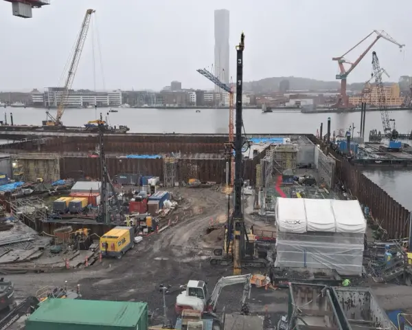 Byggarbetsplats vid vattnet med kranar, grävmaskiner och byggmaterial. Arbetet med att underminera marken pågår.