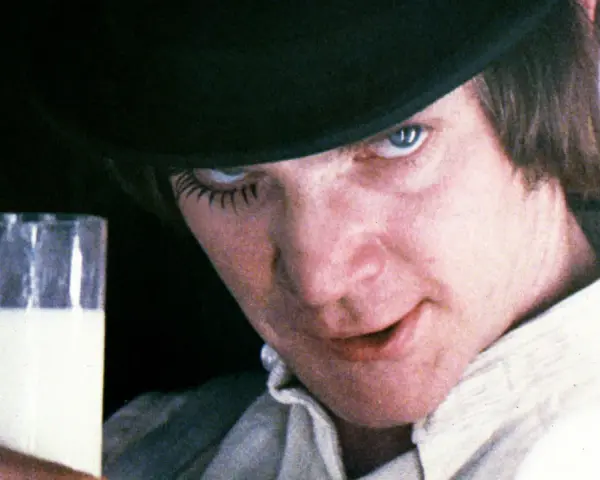 En ung man med hatt och falska ögonfransar håller ett glas mjölk. Vem skrev A Clockwork Orange? Anthony Burgess.