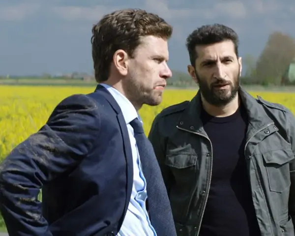 Två män står i en gul rapsåker. Den ena bär kostym, den andra en militärjacka. Scen från avdelning q film.