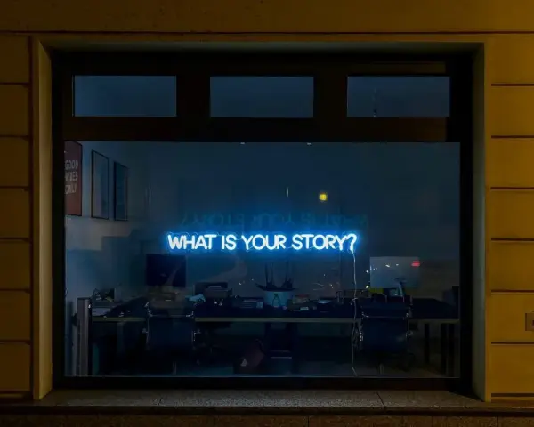 Blå neontext "WHAT IS YOUR STORY?" lyser i ett fönster, som en inbjudan till en skrivarkurs online.