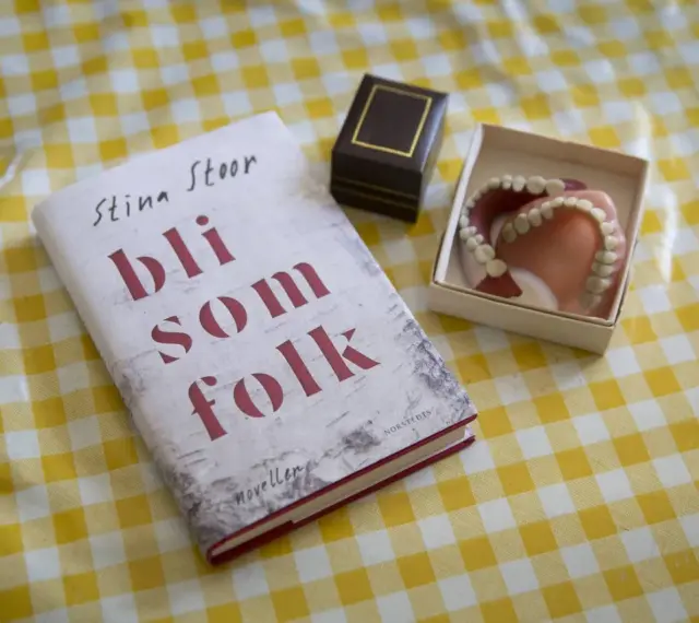 Boken "Bli som folk" av Stina Stoor ligger på ett gult rutigt bord bredvid ett par proteser.