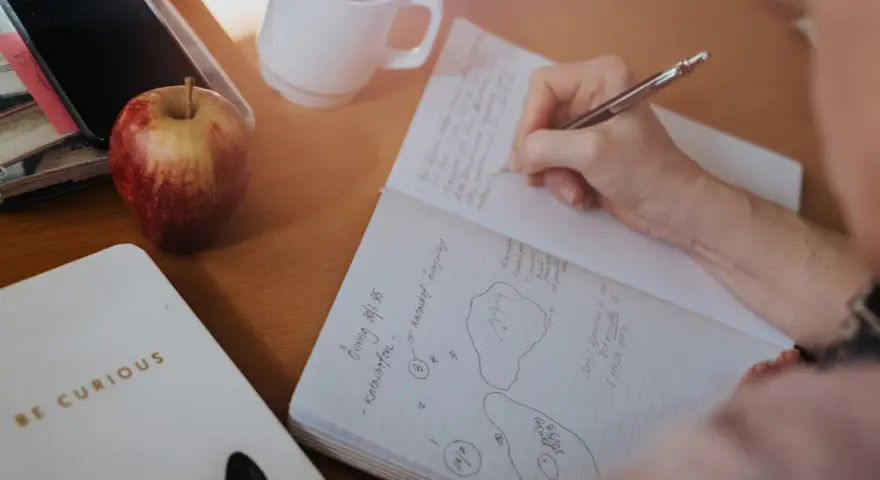 En hand skriver i ett anteckningsblock. Bredvid ligger en mobil, ett äpple och en mugg. Tidningen skriva är en del av en anteckning.