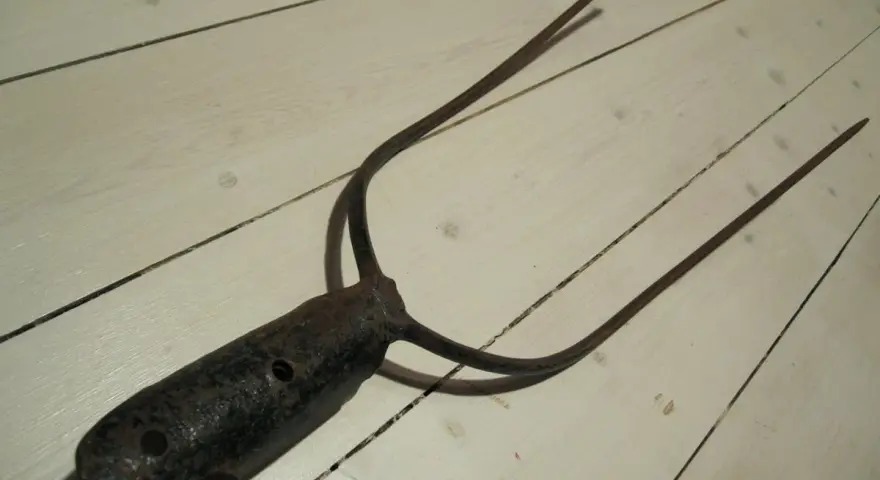 A rusty, old pitchfork with three tines lies on a white wooden floor.
