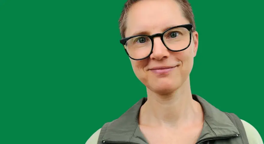 En kvinna med glasögon ler mot kameran. Hon har kortklippt hår och bär en grön väst. Bilden ger informerande text.