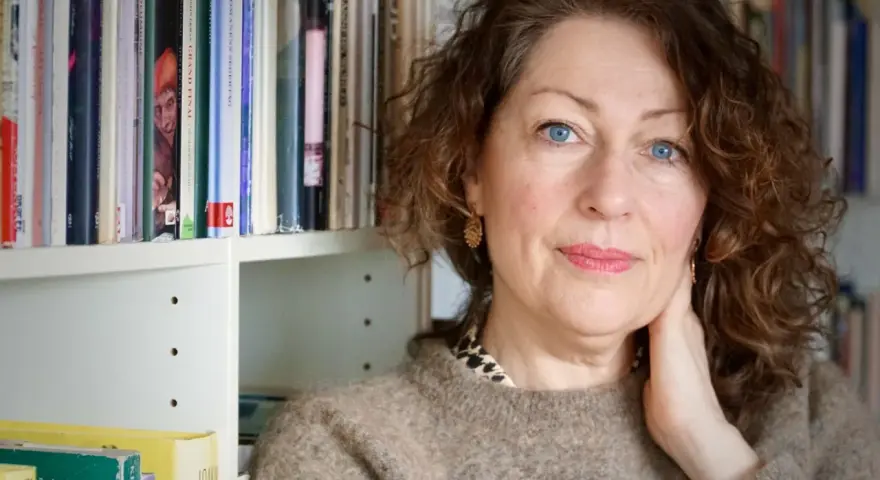 Elisabeth Åsbrink, med blå ögon och lockigt hår, sitter framför en bokhylla fylld med böcker.