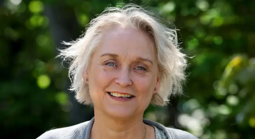 Porträtt av Karin Alvtegen med blont hår och blå ögon, leende utomhus mot en grönskande bakgrund.
