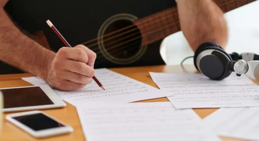 En person skriver noter med en penna, med en gitarr och hörlurar i bakgrunden. Detta är en bra inledning till en ny låt.