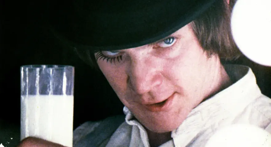 En ung man med hatt och falska ögonfransar håller ett glas mjölk. Vem skrev A Clockwork Orange? Anthony Burgess.