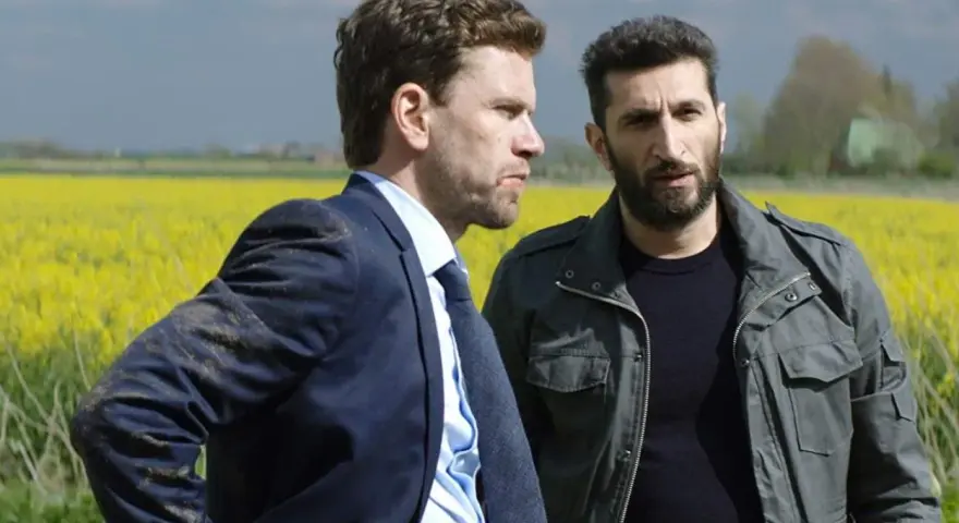 Två män står i en gul rapsåker. Den ena bär kostym, den andra en militärjacka. Scen från avdelning q film.