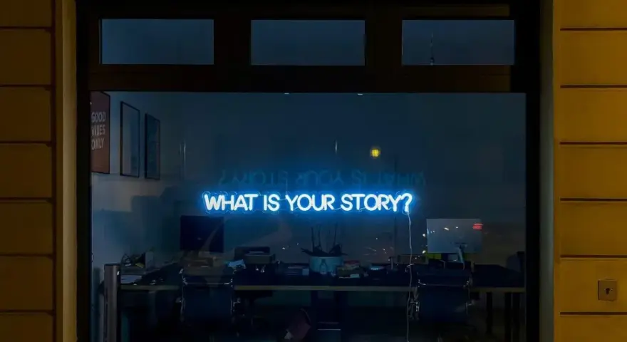 Blå neontext "WHAT IS YOUR STORY?" lyser i ett fönster, som en inbjudan till en skrivarkurs online.