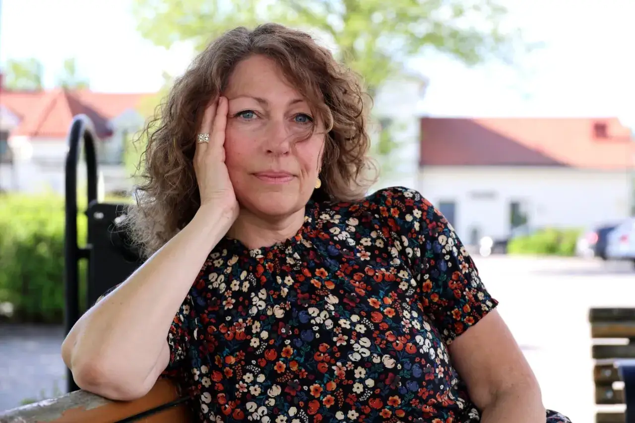 Elisabeth &Aring;sbrink sitter p&aring; en b&auml;nk med handen mot kinden, kl&auml;dd i en blommig kl&auml;nning.